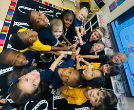 Group of children in matching uniforms forming a circle on a carpet with letters, holding hands.