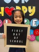 A young girl holds a sign that reads "FIRST DAY OF SCHOOL" in front of a classroom wall with a colorful "HAPPY" display.