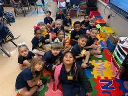 A group of children, in navy blue uniforms, sit on the floor in a classroom. A woman sits among them.