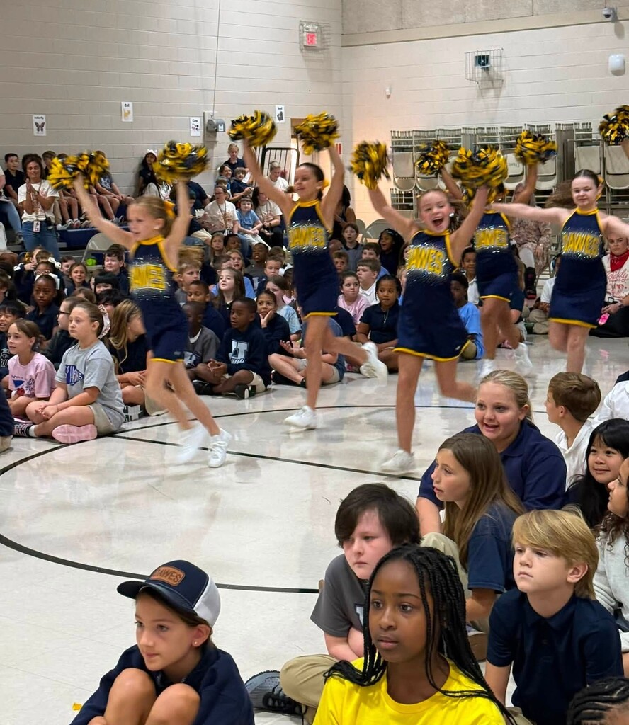 Cheerleaders run through a gym, where students are sitting on the floor