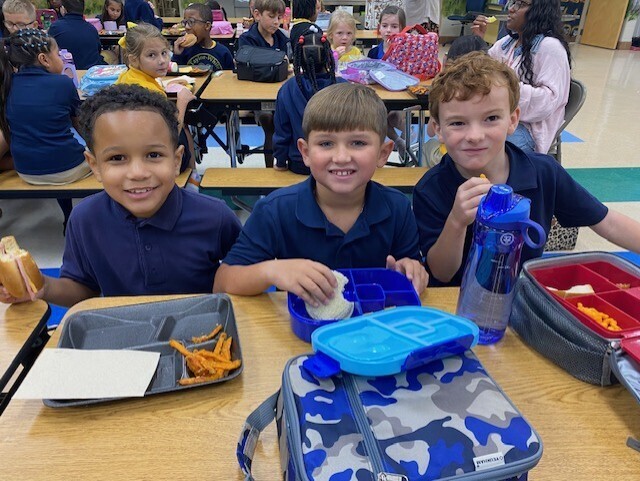 Three young boys in blue shirts sit at a table with food and drinks, with other children in the background.