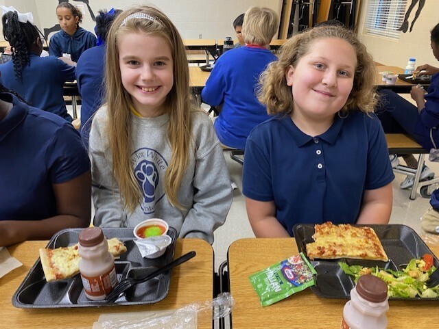 Two girls sitting at a cafeteria table with trays of food. A person in a blue shirt sits to the left, and others are in the background.
