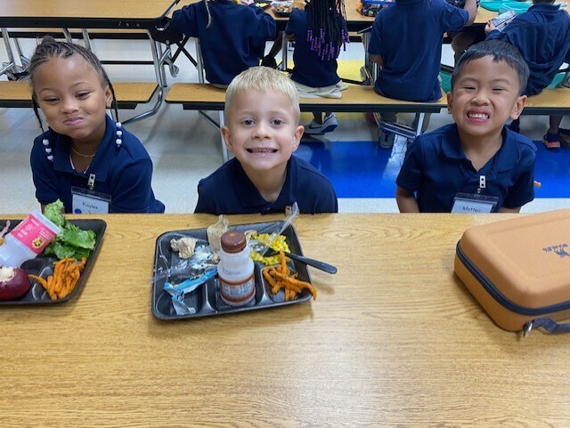 Three children in blue uniforms sit at a table with trays of food. Behind them are more students sitting at tables.