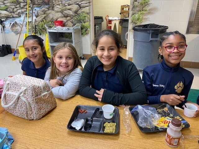 Four children at a table with a bag, cups, and plates of food in front of them.