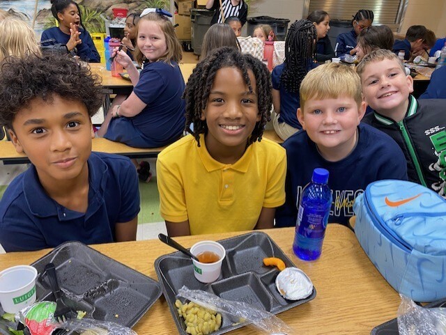 Kids at a cafeteria table with food trays, cups, and a blue backpack. Background includes other students sitting at tables.