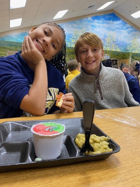 Two children sit at a cafeteria table. One holds a sandwich and smiles. Tray contains yogurt, crackers, and a spoon.