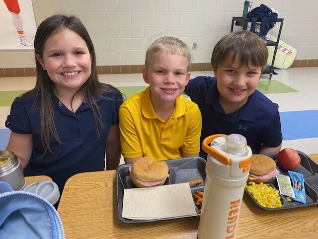 Three children, two boys and a girl, sit at a table with a tray of food, smiling.