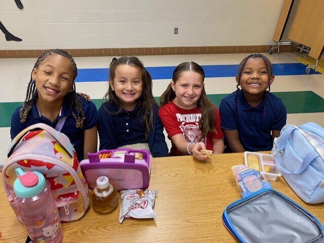 Four children at a desk with backpacks, lunch boxes, and various items. Brightly lit room with blue walls and a white floor.