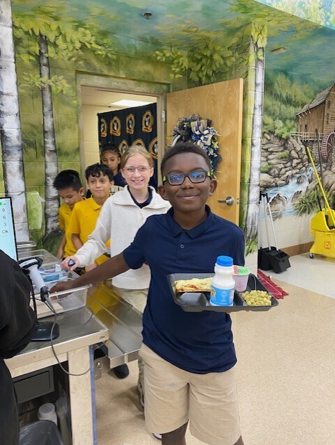 A young boy holds a tray with food in front of a mural. A girl stands behind him.
