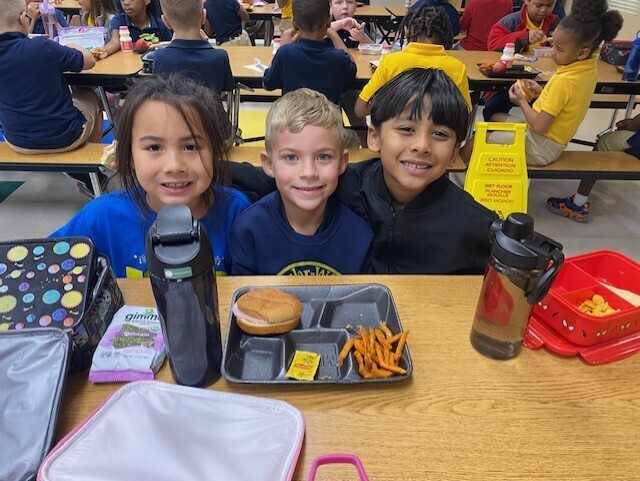 Three children sitting at a table in a classroom, smiling and posing for a picture.