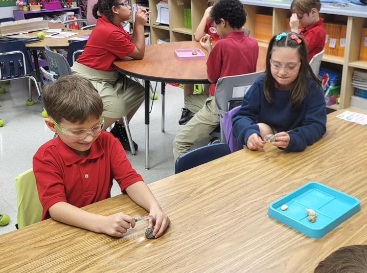 miss Caballero’s class observing rocks with magnifying glass in their classroom sitting at their tables