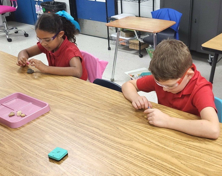 miss Caballero’s class observing rocks with magnifying glass in their classroom sitting at their tables