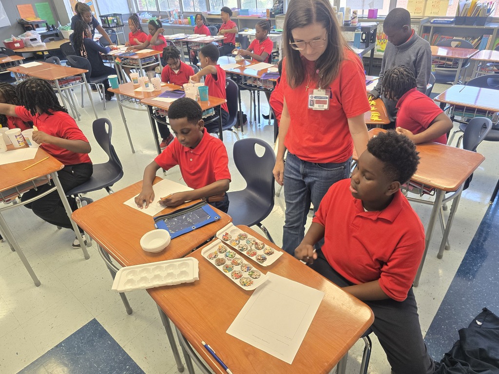 A classroom with students and a teacher. Students in red shirts sit at desks, one with a tablet.