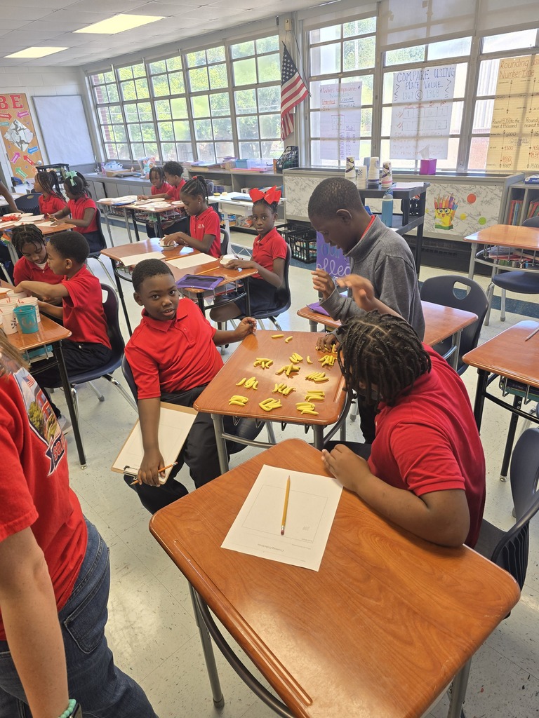 In a classroom, several students wearing red shirts sit at desks with various items. One student leans over a desk.