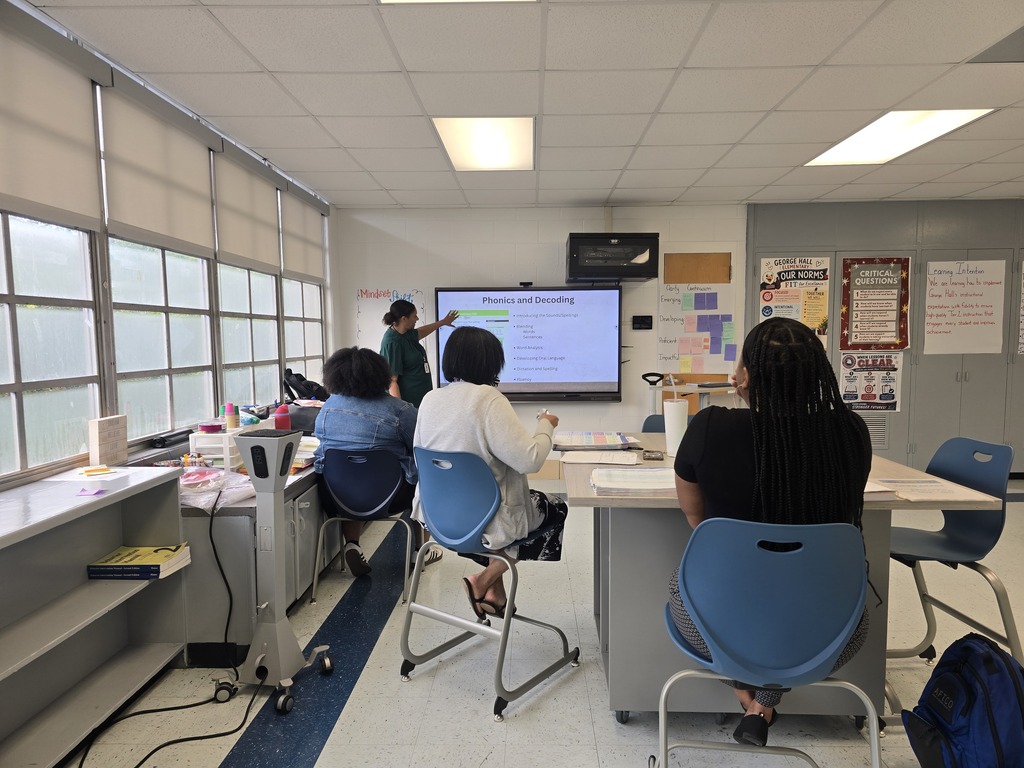 Four people sit at tables in a classroom. One stands in front of a screen and presents.