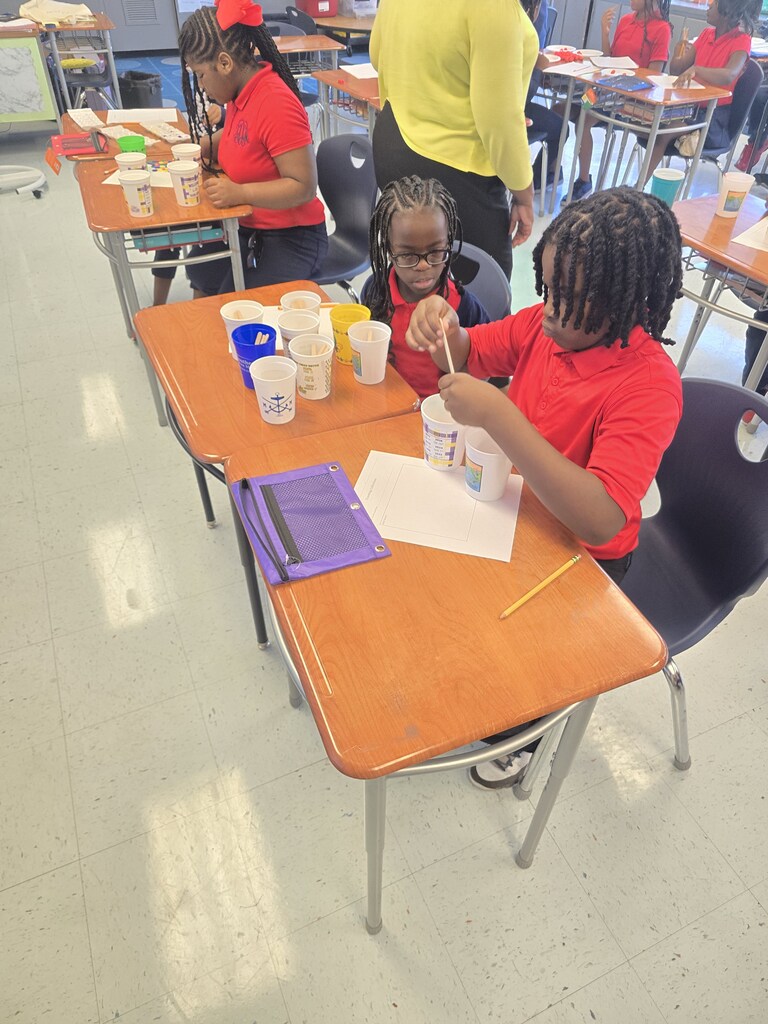 Kids in red shirts seated at desks in a classroom. One child holds a paintbrush. Cups and paper on the desks.