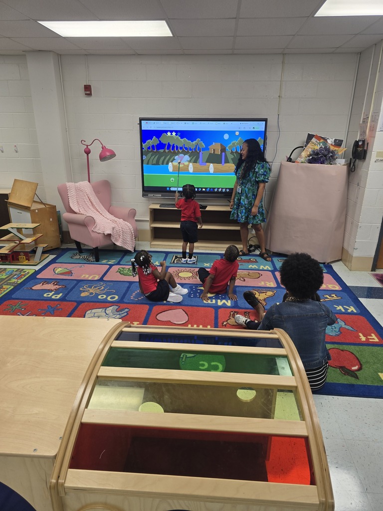 A woman stands before a TV screen in a playroom. Four children sit on the floor, watching.