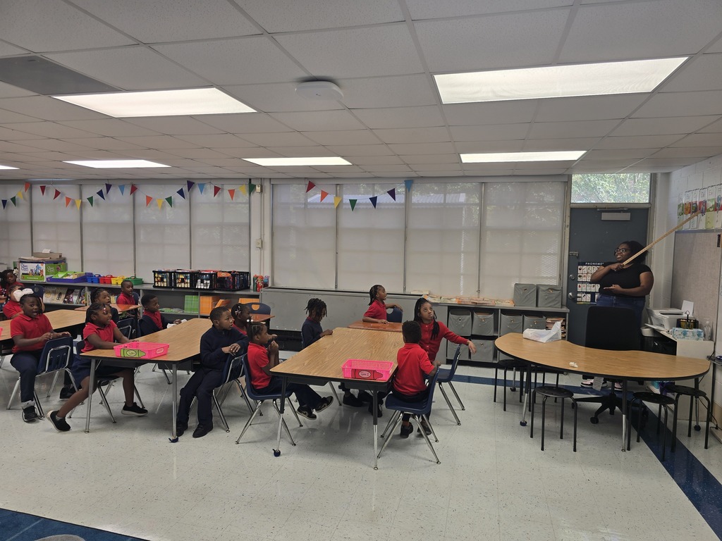 Students in red shirts seated at tables in a classroom with a teacher standing by a door.