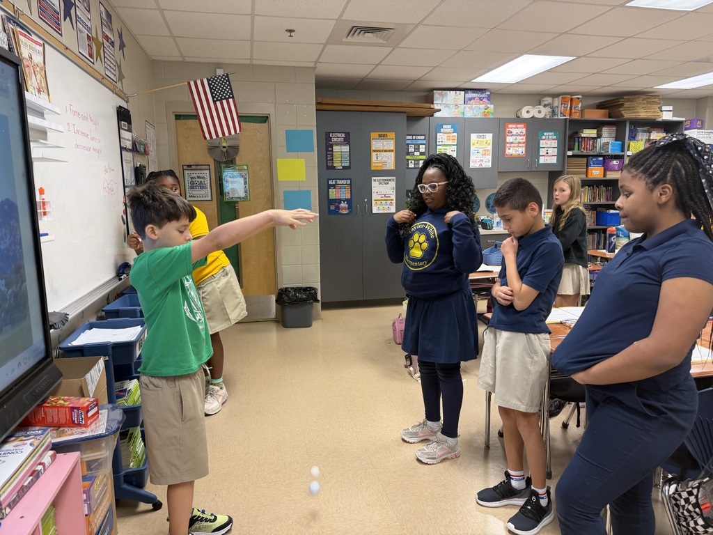 Several students in a classroom are standing and talking. A boy points at a monitor.