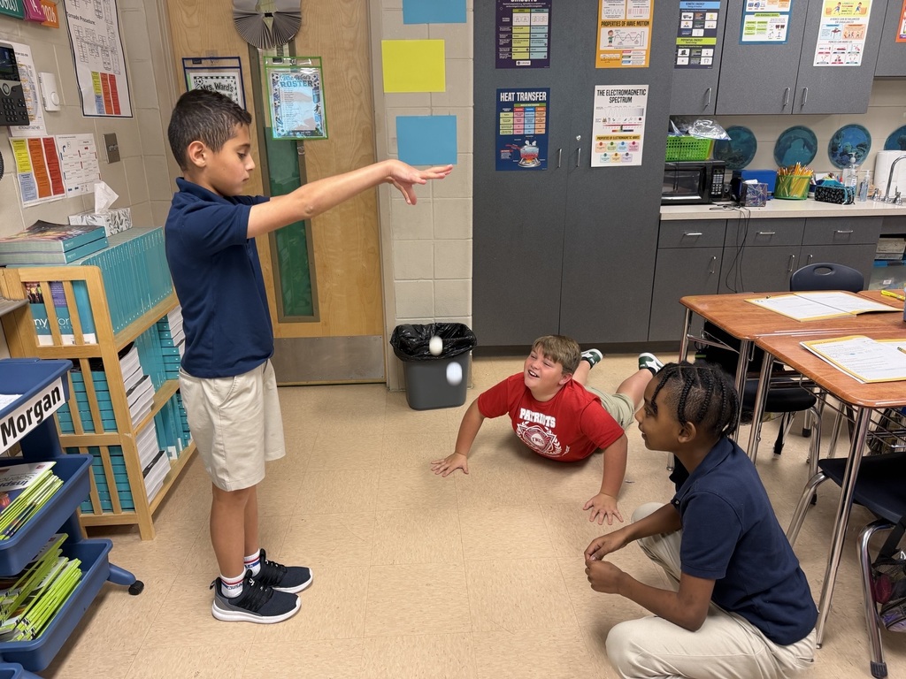 A child pointing, while another child kneels and a third child sits, in a classroom with desks, cabinets, and bulletin boards.