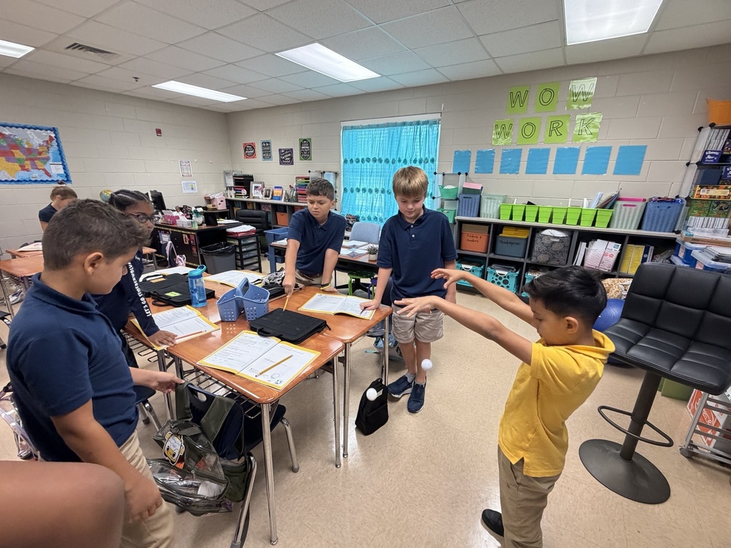 Five boys in a classroom. One holds up a hand. Desks, chairs, and a table with papers.