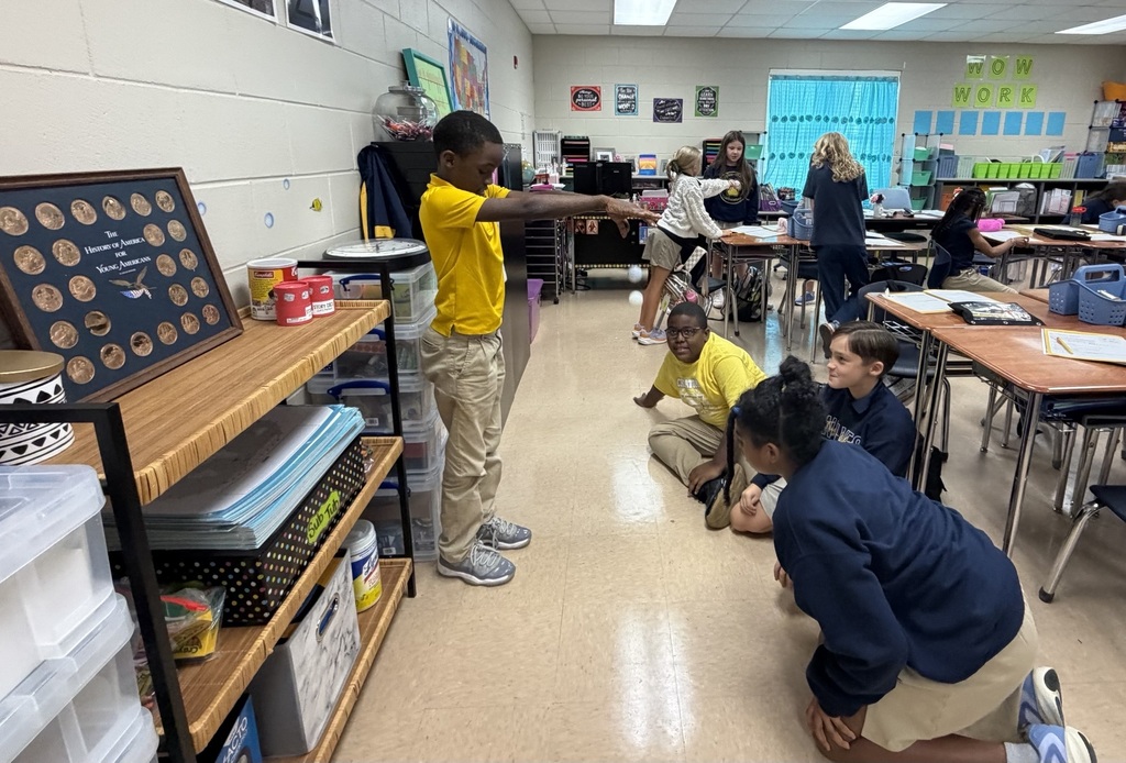 A classroom with students seated and standing; a boy in yellow points to the side. Tables, chairs, and shelves with items.