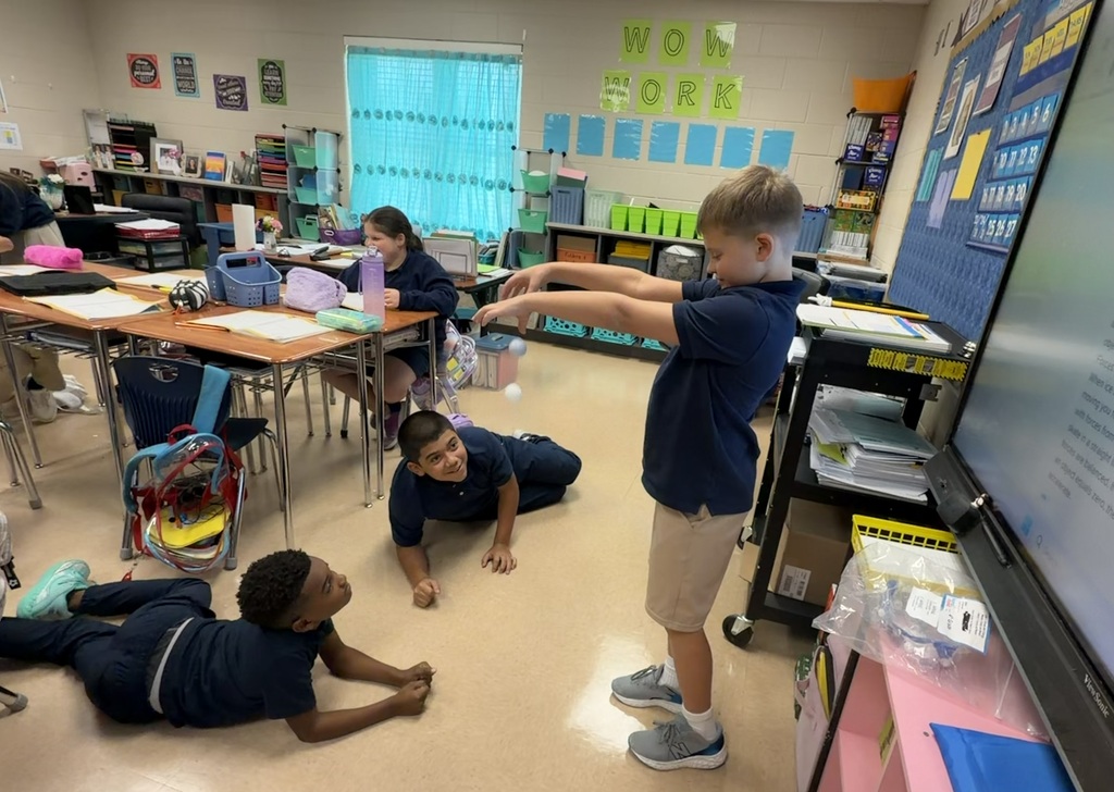 Children in a classroom. One student stands while the others are on the floor. A large board is behind them.