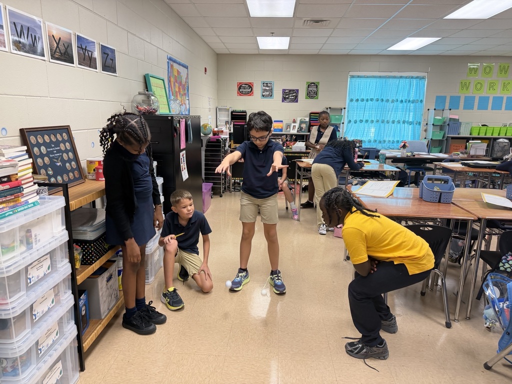 A classroom with desks and shelves. Children crouch or stand, facing each other in pairs. One boy in yellow crouches, one in blue stands, and two in black crouch.
