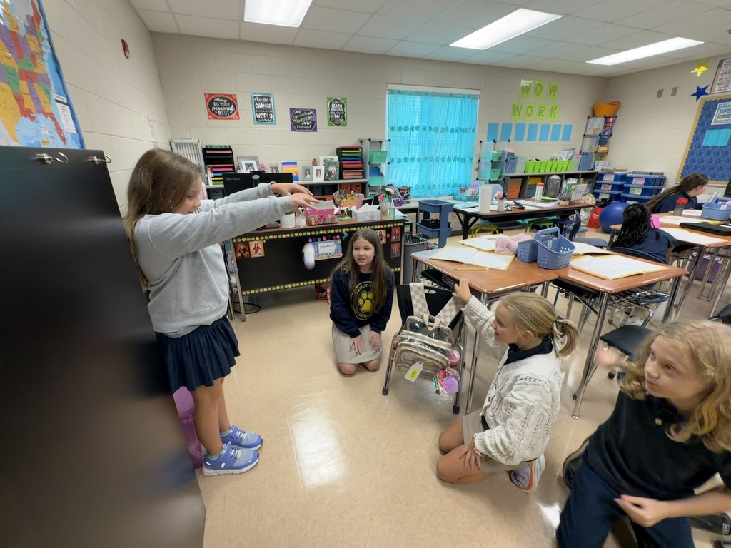 Several young students in a classroom, with one student gesturing toward a board and others seated at desks.
