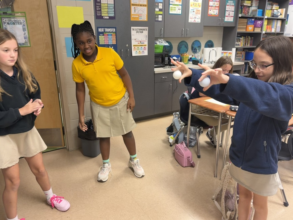 Four girls in school uniforms, two holding balls, in a classroom with a desk, trash can, and shelves.