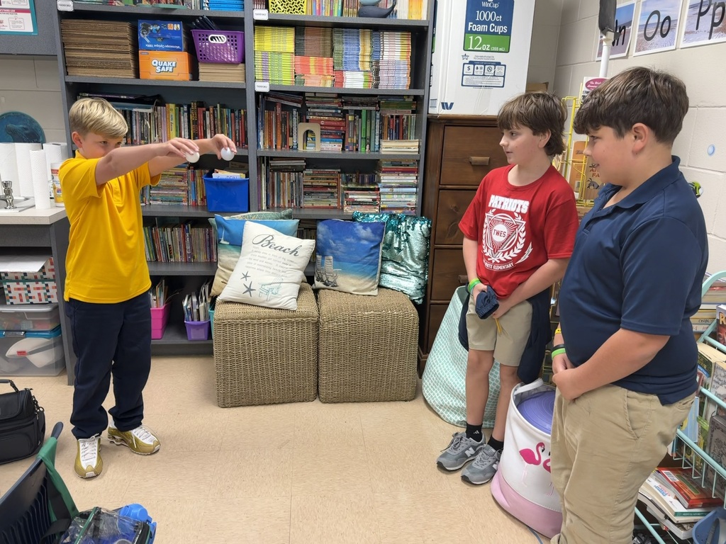 Three young boys in a classroom with bookshelves; one points to a blue object in his hand.