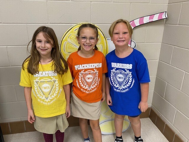 Three children stand together in a hallway, each wearing different colored shirts. The background includes a wall with white tiles.