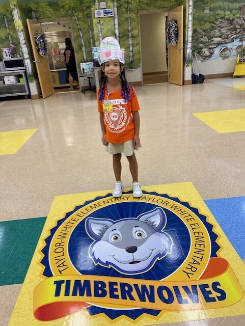 Child in orange shirt and shorts stands on Timberwolves school logo on the floor. Behind the child, a wall with doors.