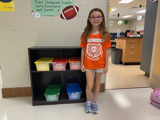 A girl in an orange shirt stands next to a black bookshelf with colorful bins in a classroom.