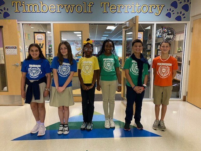 Five children stand on a paw print, each wearing different colored shirts. Behind them is a building with glass doors.