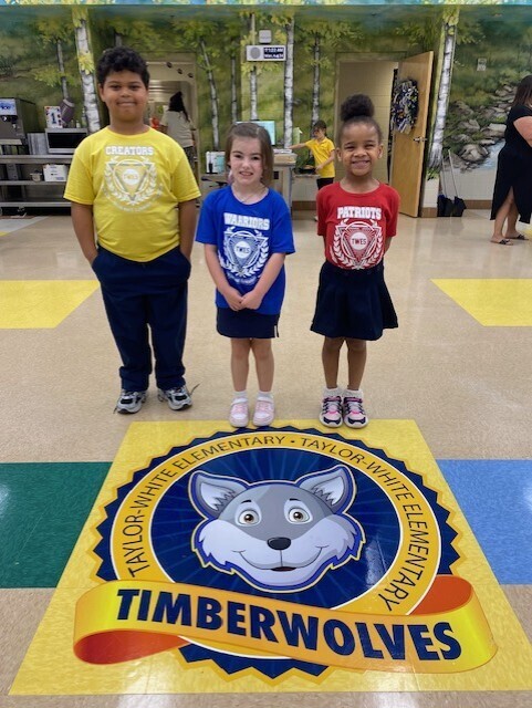 Three children stand on a floor with a Timberwolves logo. They wear school uniforms. The background shows a door and a mural.