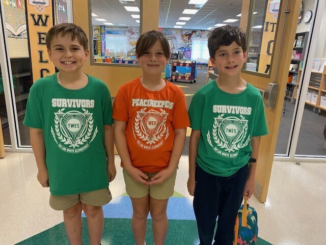Three children in matching green t-shirts, one wearing orange, stand in a hallway. Behind them is a glass door.