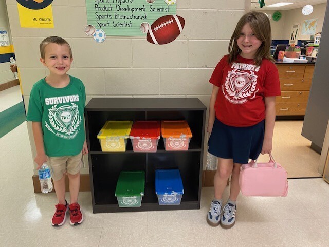 Two children stand next to a colorful shelf. One holds a water bottle, the other a pink bag.