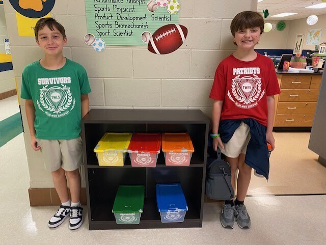 Two boys stand by a shelf with bins in various colors. One wears a green shirt and the other a red one.