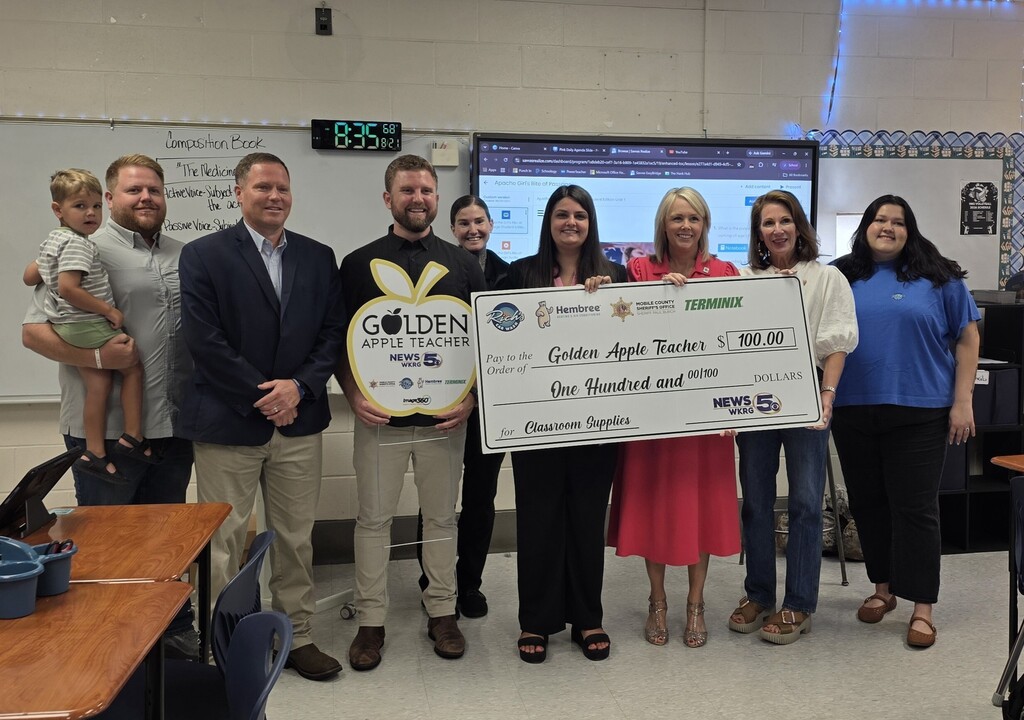 A teacher holds an oversized check while posing with her principal, family members, fellow teachers and Devon Walsh of WKRG