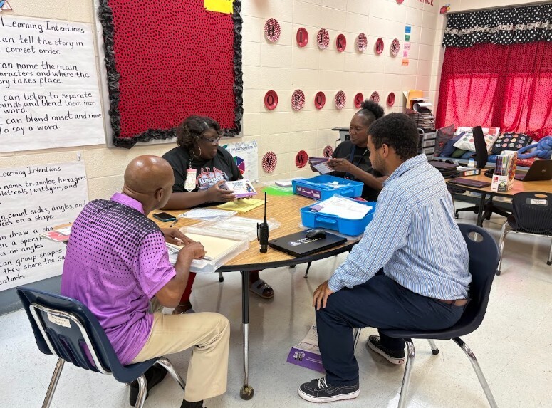 A group of adults sit around a table in a classroom. Papers, a tablet, and a pen are on the table.