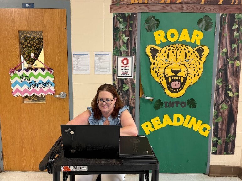 A woman wearing glasses sits at a table with a laptop, in front of a door with a green banner reading "ROAR INTO READING."