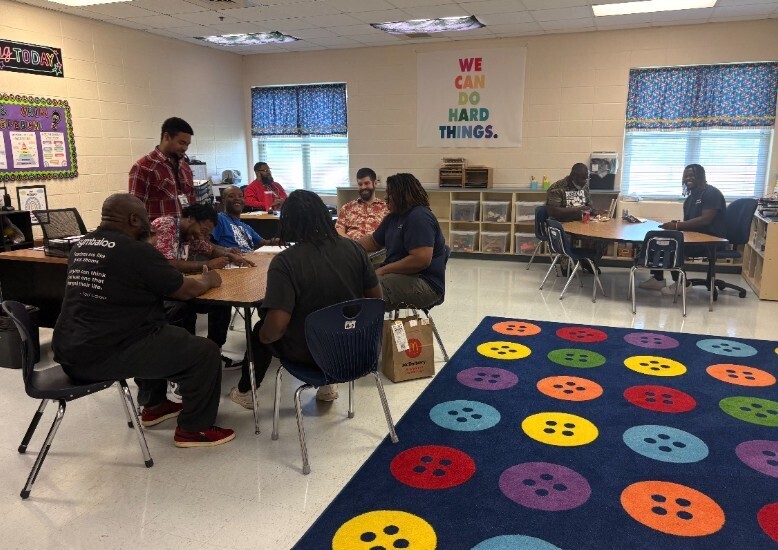 People sitting at tables in a classroom. Bright colorful rug. Poster on wall says, "We can do hard things."