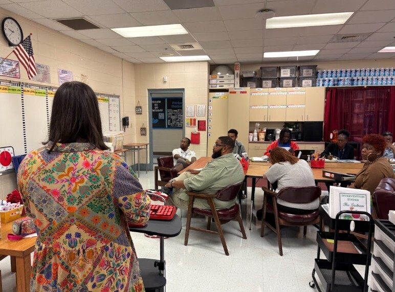 A classroom with people sitting around tables. A person stands in front of a table.