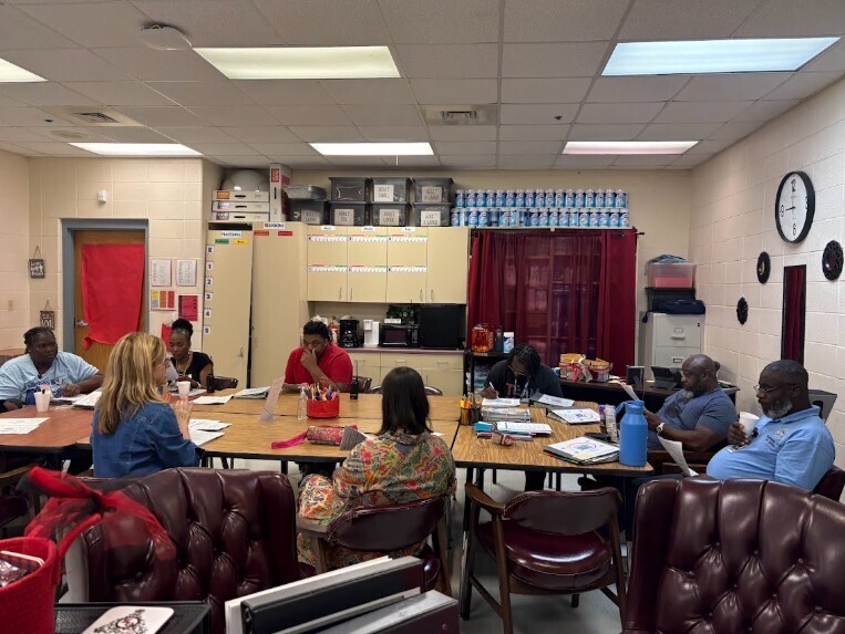 A group of people are sitting around a table in a classroom with tables and chairs.