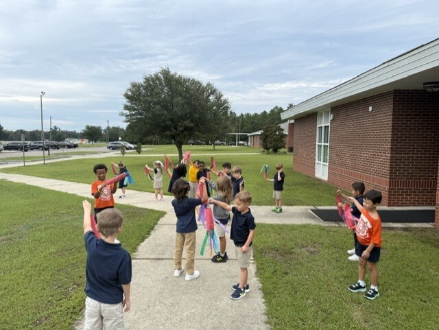 A group of children in blue and orange shirts holding streamers stand in a line on a sidewalk.