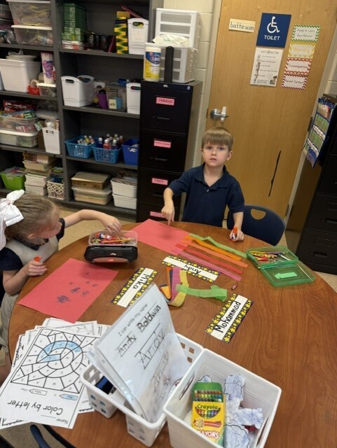 Two children sit at a table with art supplies, including papers, scissors, and crayons. Behind them is a door with an accessible symbol and shelves with boxes and bins.
