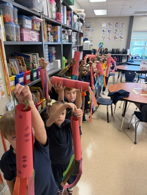 A line of children in a classroom, each holding a pink tube. Shelves with books and storage boxes in background.