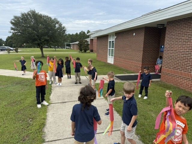 Children in blue shirts and shorts holding colorful ribbons, walking on a pathway in front of a brick building.