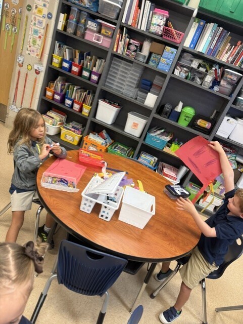 Three children seated around a round table with books and craft supplies. One child holds up a red paper.